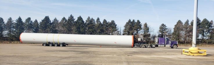 A wind tower section on a trailer in a paved lot lined with pines