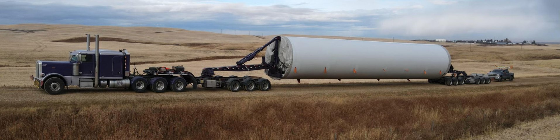 Purple FT&L Specialized Peterbilt hauling a white wind tower section across open prairie under a wide sky