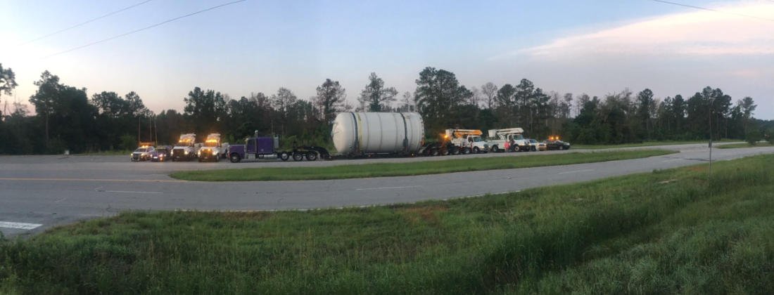 An FT&L crew and escort vehicles staged along a rural highway at dusk beside an over-dimensional load