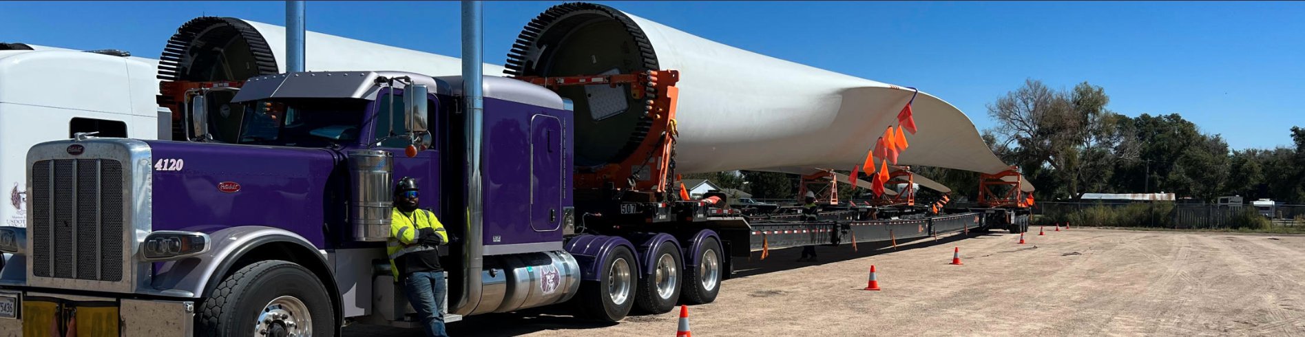 A purple FT&L Peterbilt hauling a white wind tower section along an open highway